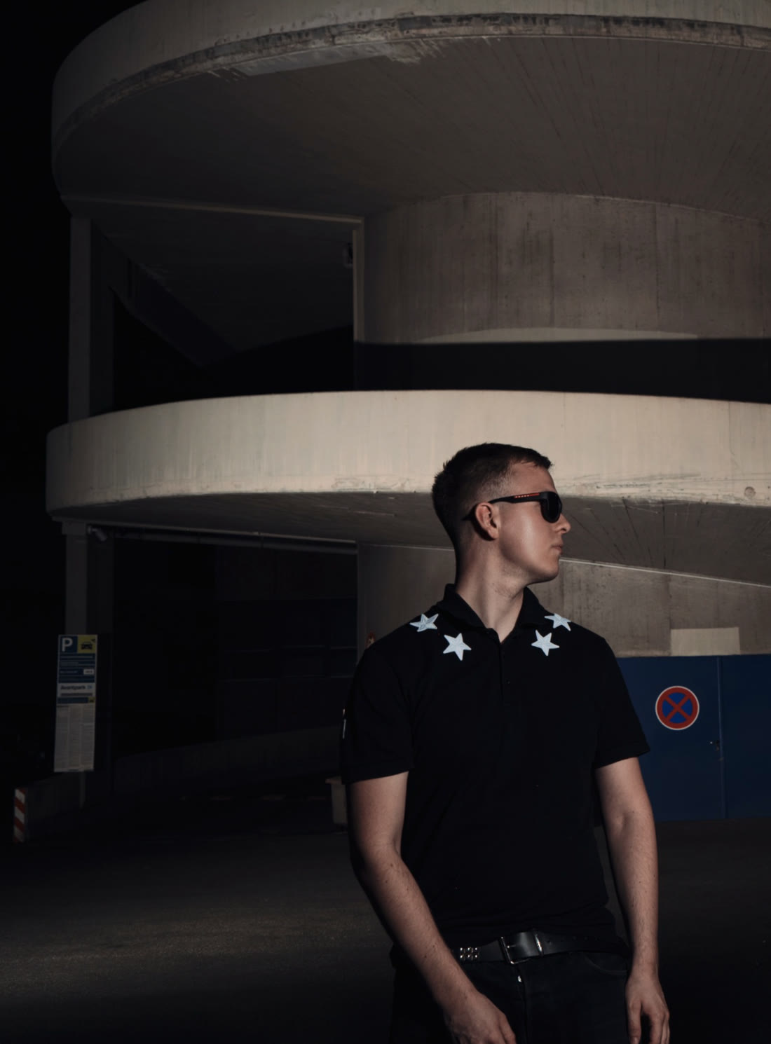 madein2k photographed at night in front of a spiral parking garage, wearing a star-collar polo and sunglasses.